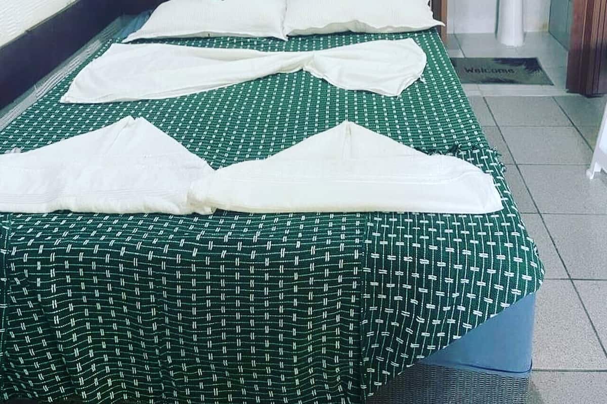 Una cama bien hecha con una colcha verde y blanca a cuadros, almohadas blancas, una puerta de madera y un baño con lavamanos y espejo.