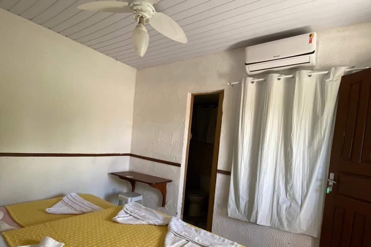 Habitación de hotel con dos camas, ventilador de techo, equipo de aire acondicionado, una mesita y una ventana con cortinas.
