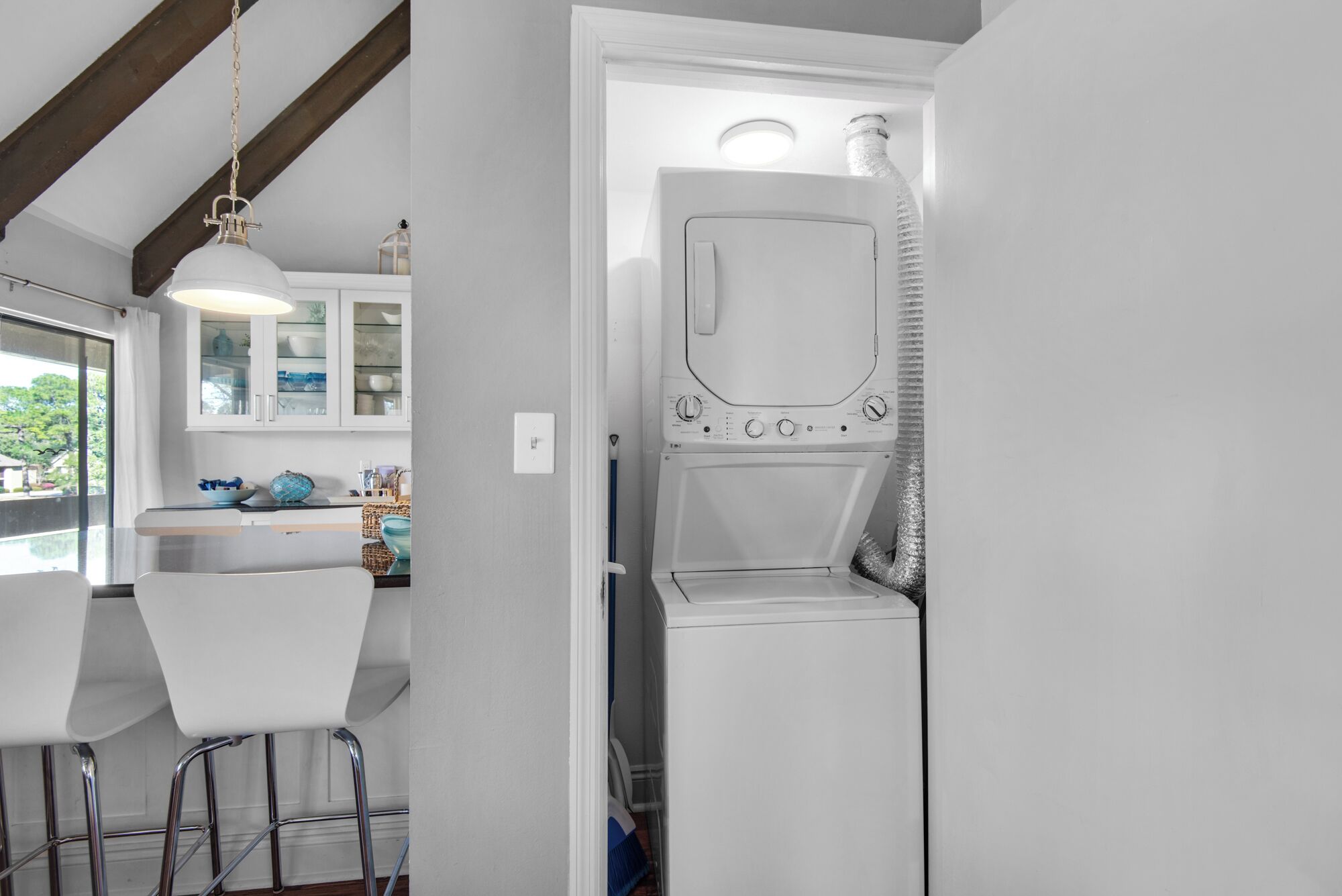 Une cuisine compacte équipée d’un lave-linge et d’un sèche-linge intégrés, d’un tabouret de bar blanc et d’un luminaire suspendu.