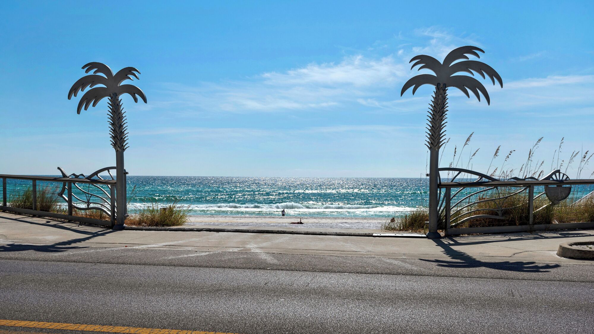 A coastal road with metal palm tree sculptures on either side, leading to a beach with waves crashing onto the shore.