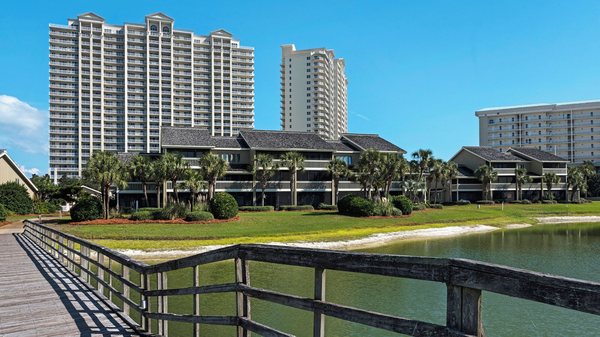 A waterfront hotel with multiple high-rise buildings in the background, a wooden boardwalk leading to the water, and well-maintained landscaping.