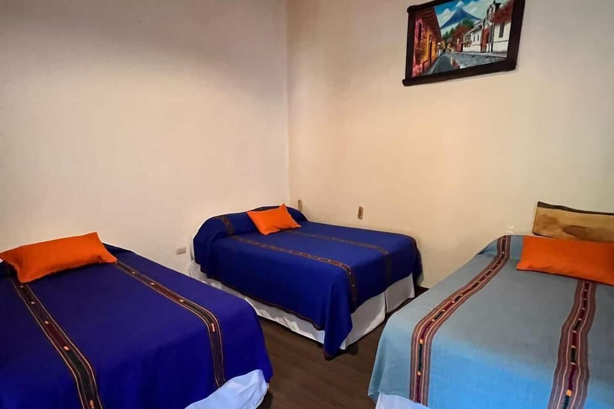 Una habitación con tres camas, cada una con un edredón de diferente color. Hay un cuadro en la pared sobre las camas.