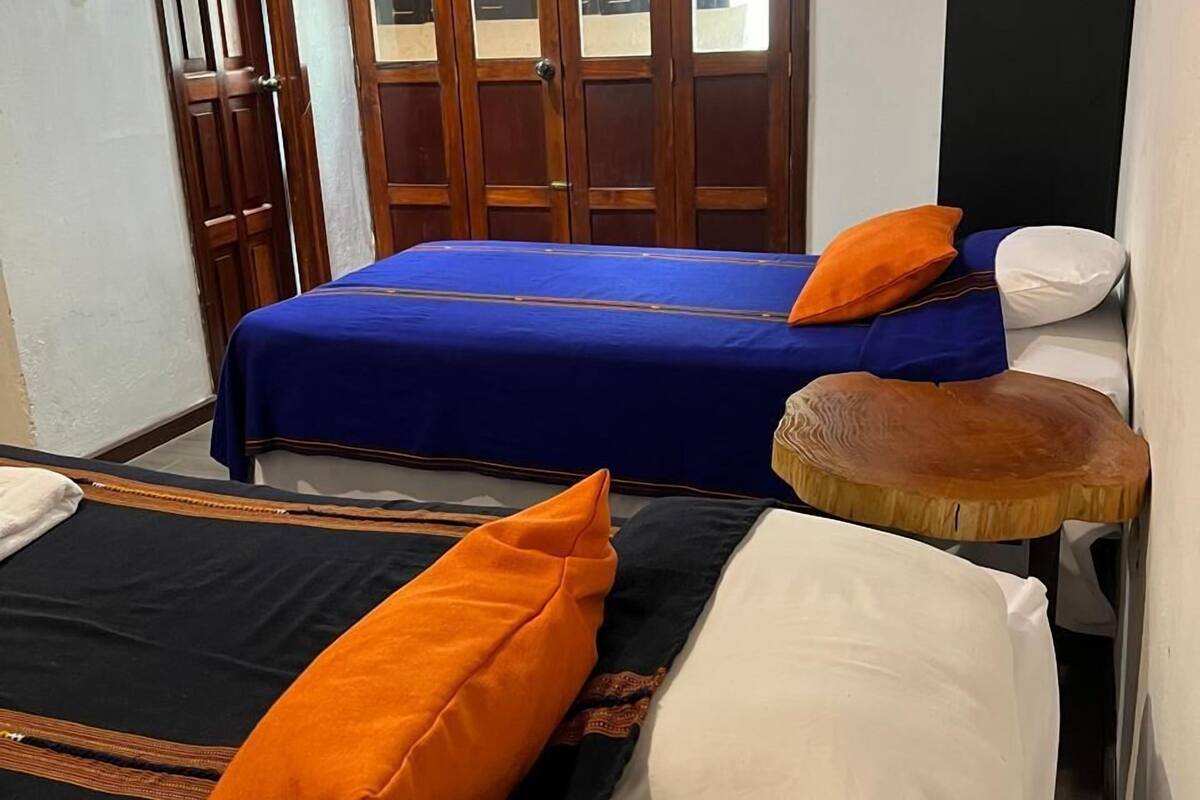 Una habitación con dos camas, una con colcha azul y la otra con colcha negra. Hay almohadas naranjas en ambas camas. Un taburete de madera está colocado junto a las camas.