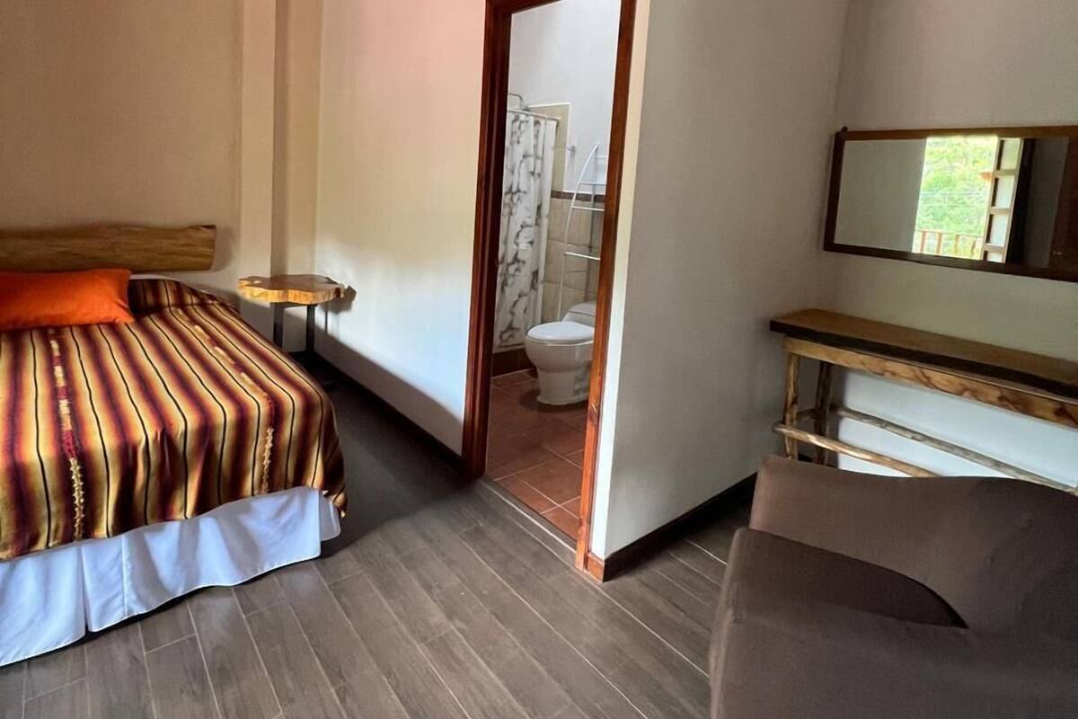 Un dormitorio con cama, una mesita de noche, una silla, un espejo y vista al baño.
