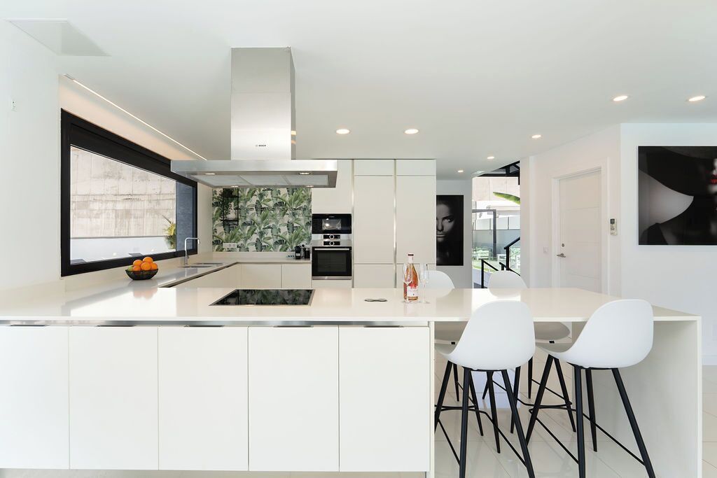 Une cuisine moderne avec des meubles blancs, un îlot central doté de tabourets et une grande fenêtre laissant entrer la lumière naturelle.