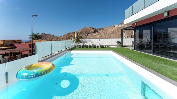 A modern pool area with clear blue water, a floating ring, and a view of a rocky hillside.