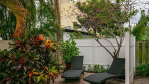 Une terrasse avec deux fauteuils de jardin en osier, une clôture blanche et diverses plantes tropicales.