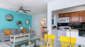 Ein modernes Wohnzimmer mit Sofa, Couchtisch und Sesseln, eine Küche mit Holzschränken und Geräten sowie ein Deckenventilator.