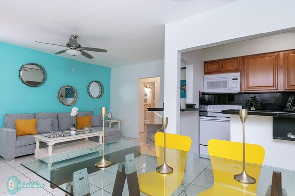 A modern living room with a sofa, coffee table, and chairs, a kitchen with wooden cabinets and appliances, and a ceiling fan.