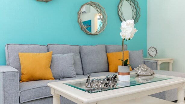 Ein Wohnzimmer mit grauem Sofa, gelben Kissen, einem Glastisch mit Namensschild und einer weißen Orchidee im weißen Topf.
