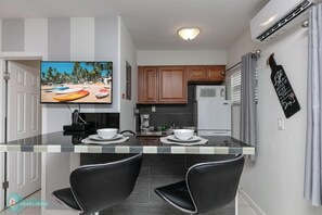 A modern kitchen with a dining area, featuring a TV, two black chairs, a white table, and a kitchen island. - 1bd 1bth, PYT, Airport, 17th Street (Fort Lauderdale)