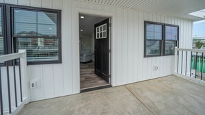 A covered porch with a white exterior, a door leading to an interior room, and a view of a residential area in the background.