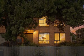 Uma casa de dois andares com fachada de tijolos, janelas amplas e cerca de piquete branca.