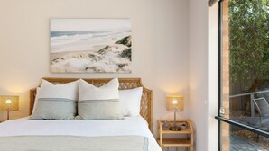 Ett sovrum med en stor säng, två sänglampor, en träpanel som utgör sänggaveln, ett fönster med utsikt över en strand och ett litet träbord vid sängen med en lampa och en liten prydnadsfigur.