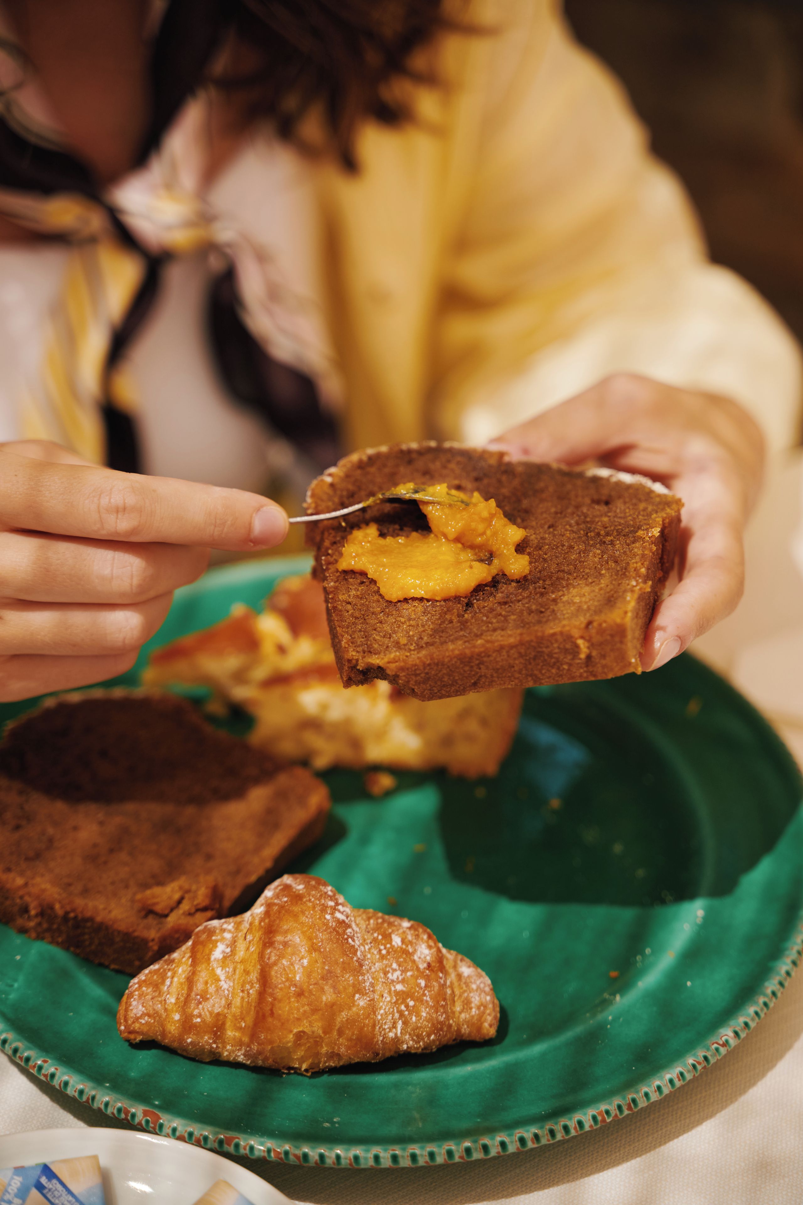 Une personne tenant un muffin avec une cuillère, une assiette verte sur laquelle se trouve une pâtisserie, et une vue partielle d’une pièce.