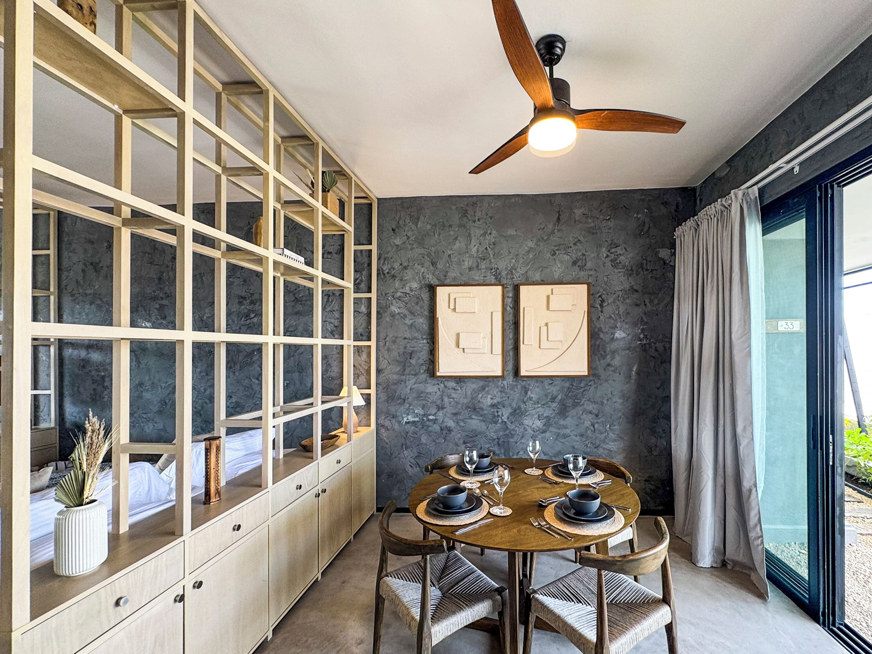 Un coin repas avec une table ronde pour quatre personnes, des chaises en bois, un ventilateur de plafond et une vue à travers une porte vitrée.