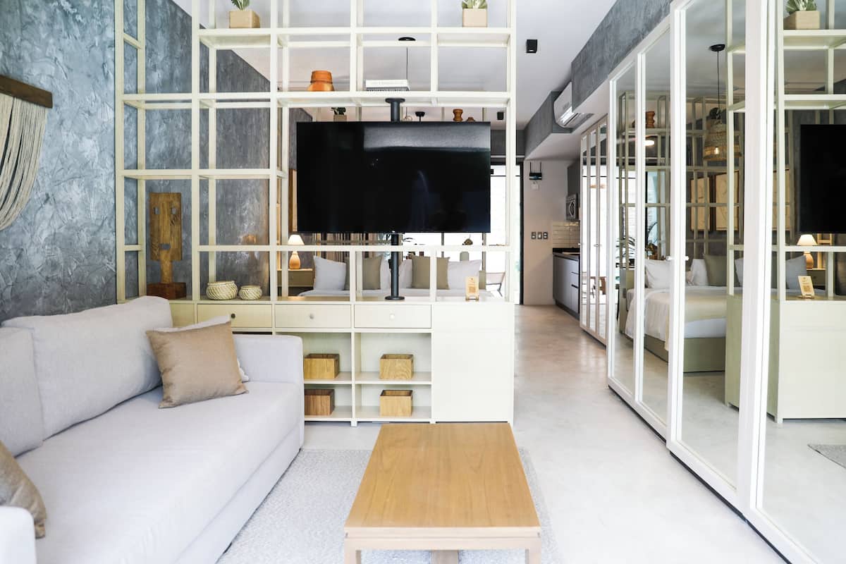 A modern living room with a sofa, a coffee table, and a TV. There is a white shelving unit with decorative items and a kitchen area visible through an open door.
