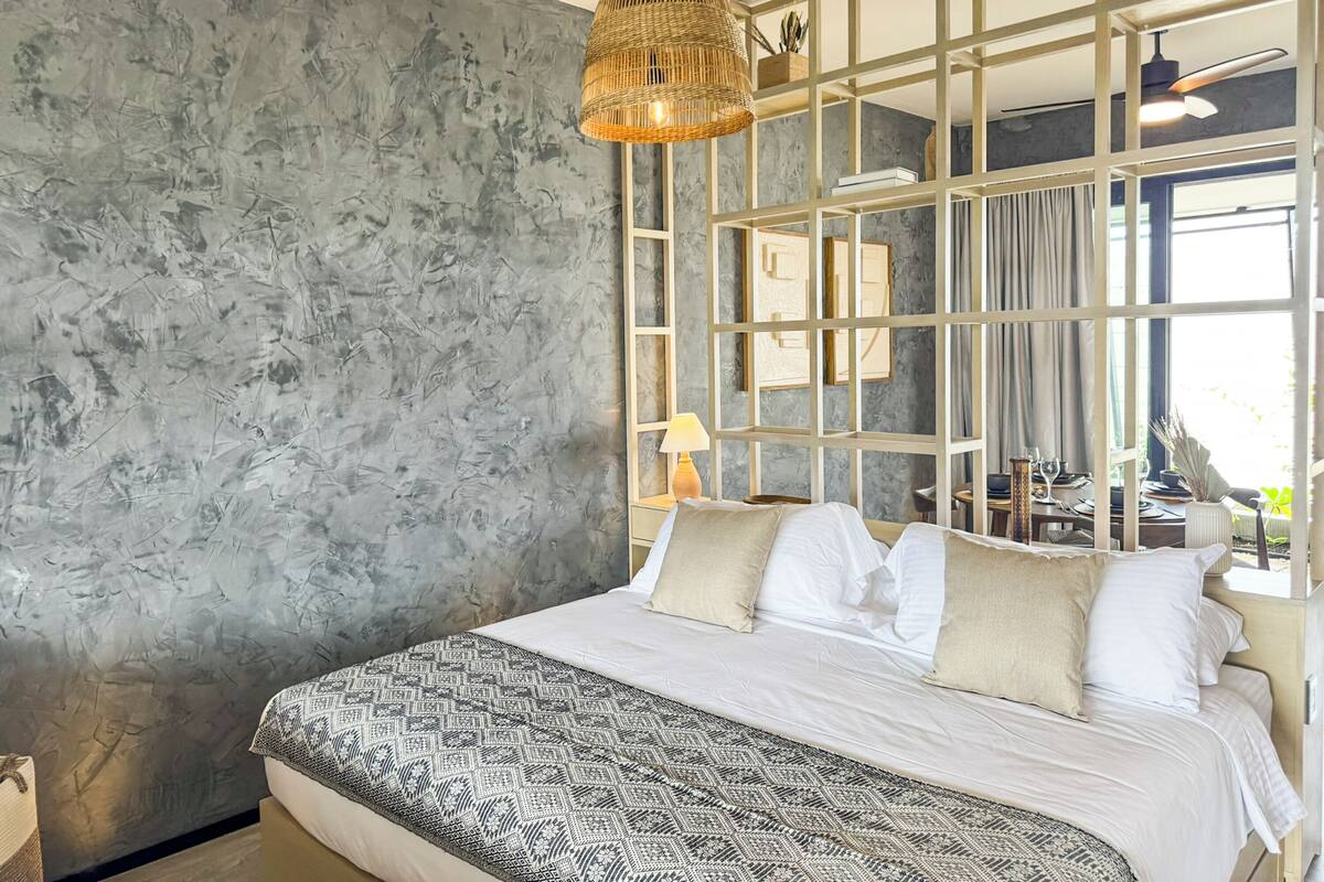 A bedroom with a large bed, a textured gray wall, a woven light fixture, and a view of a dining area through a glass partition.
