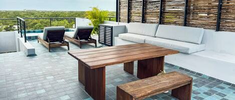 A rooftop terrace with a wooden table and benches, a white sofa, and a view of the cityscape.