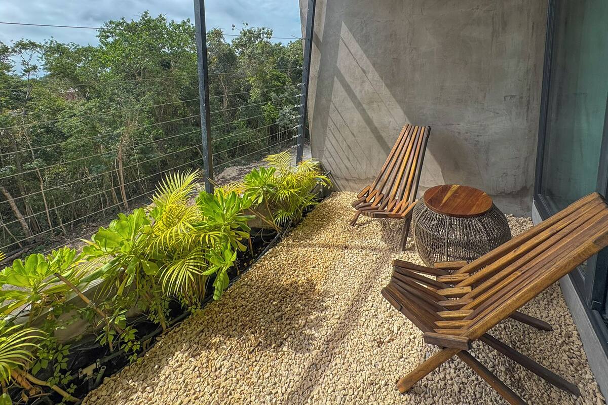 A balcony with two wooden chairs and a round wooden table, surrounded by gravel and green plants.
