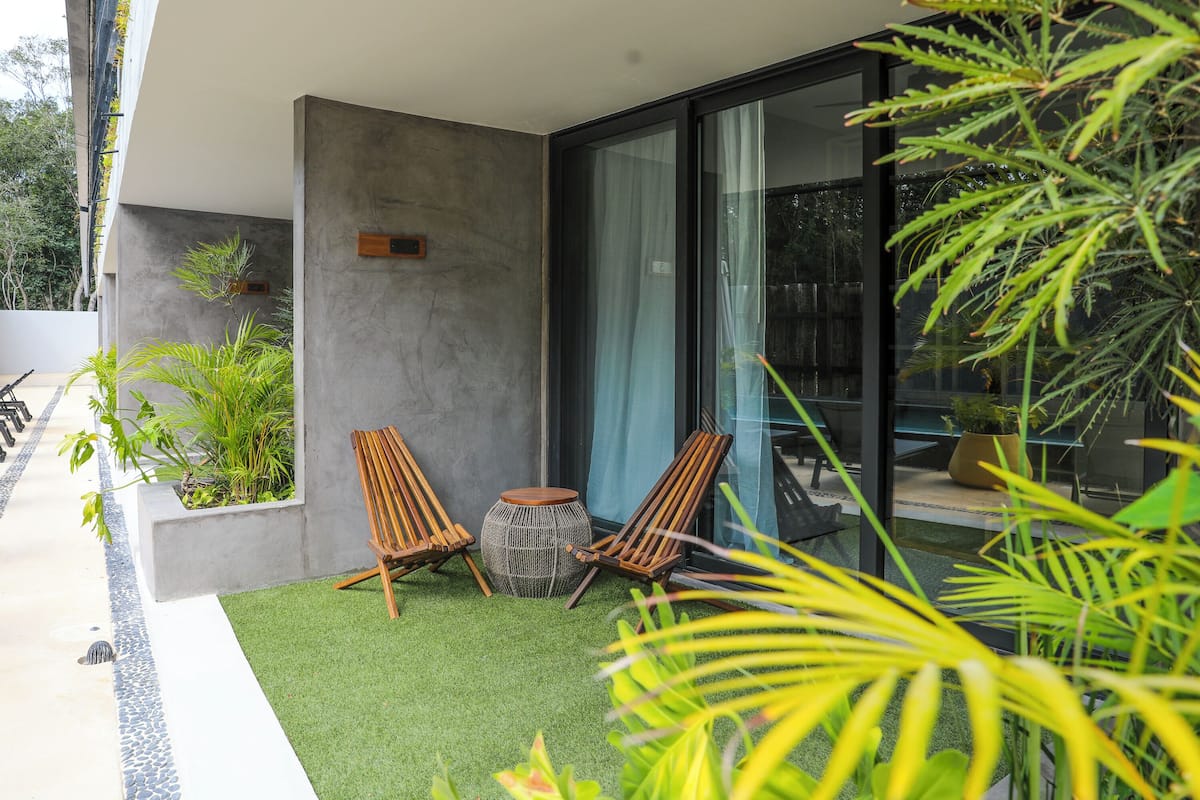 A modern outdoor seating area with two wooden chairs and a woven stool, surrounded by greenery and a glass door leading to a patio.