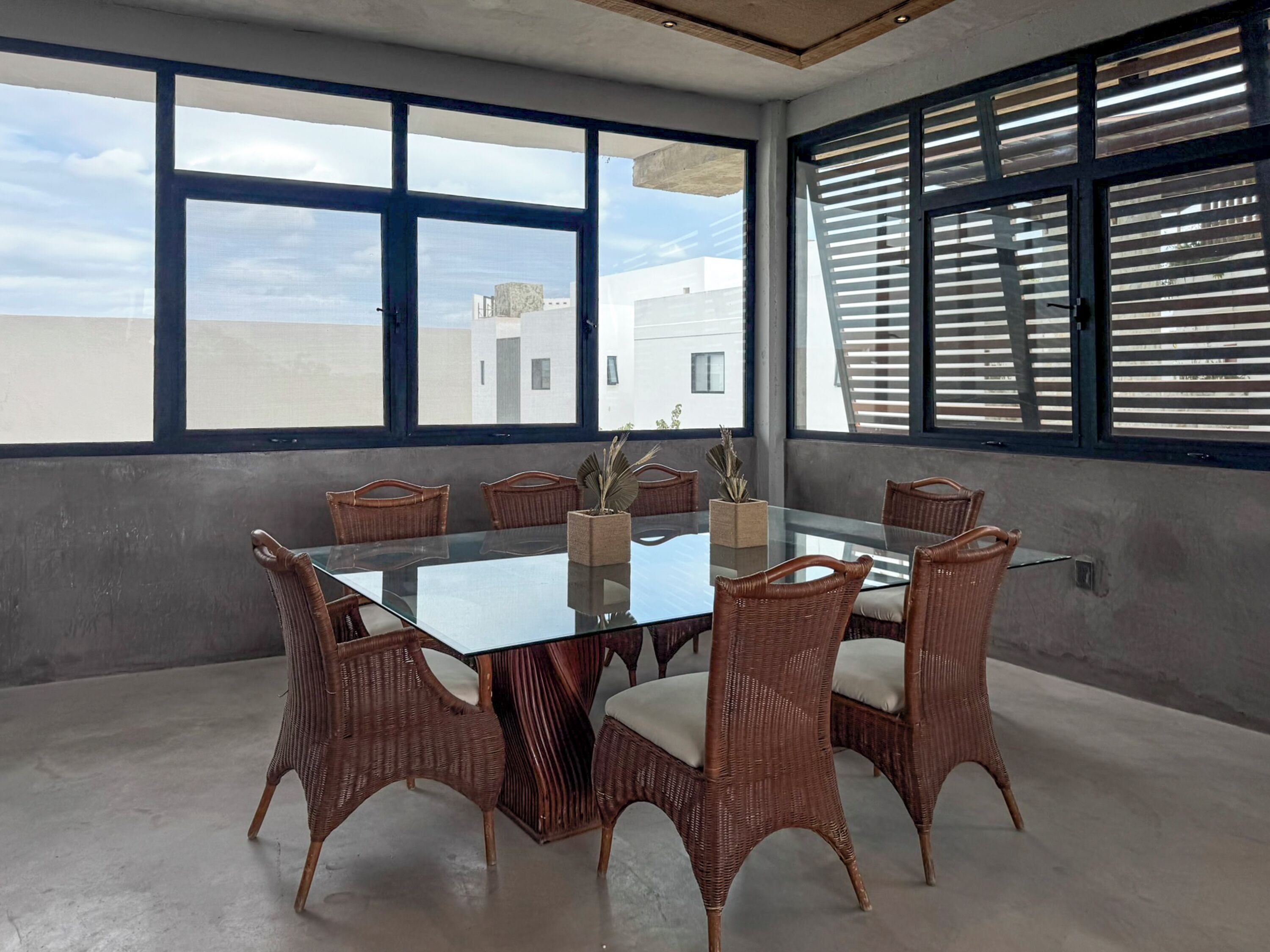 A modern meeting room with a glass table and wicker chairs, large windows with blinds, and a view of buildings outside.