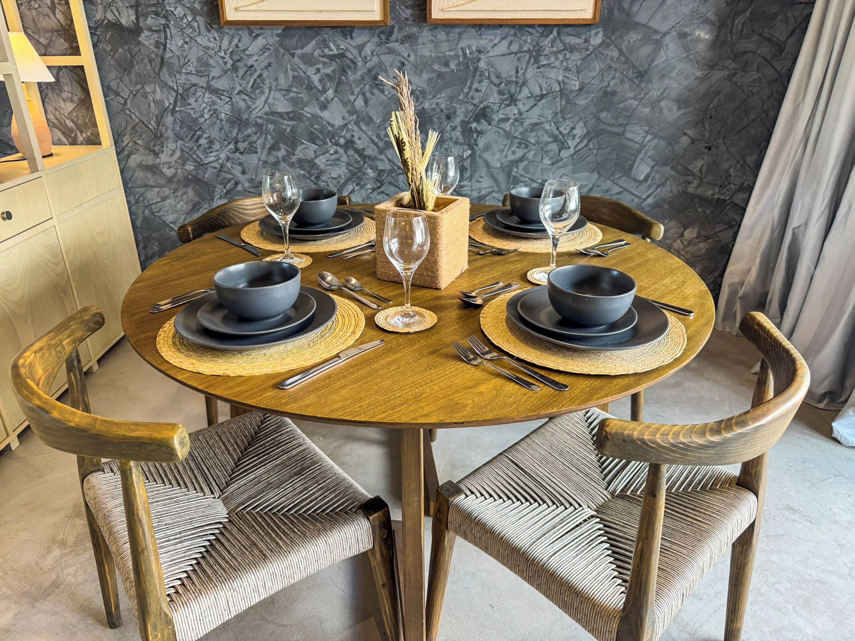 A round wooden dining table with black bowls and silverware, surrounded by wicker chairs.