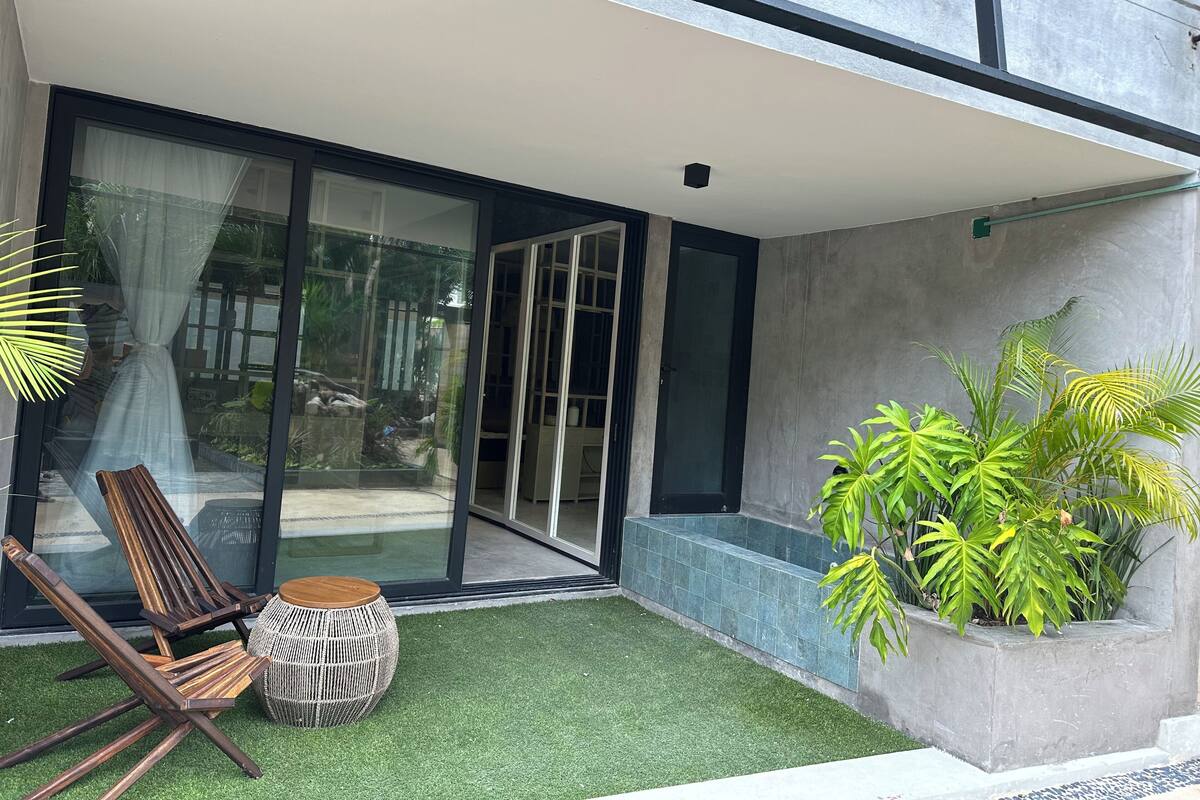 A modern outdoor seating area with a glass door leading to a patio, surrounded by greenery and a concrete planter.
