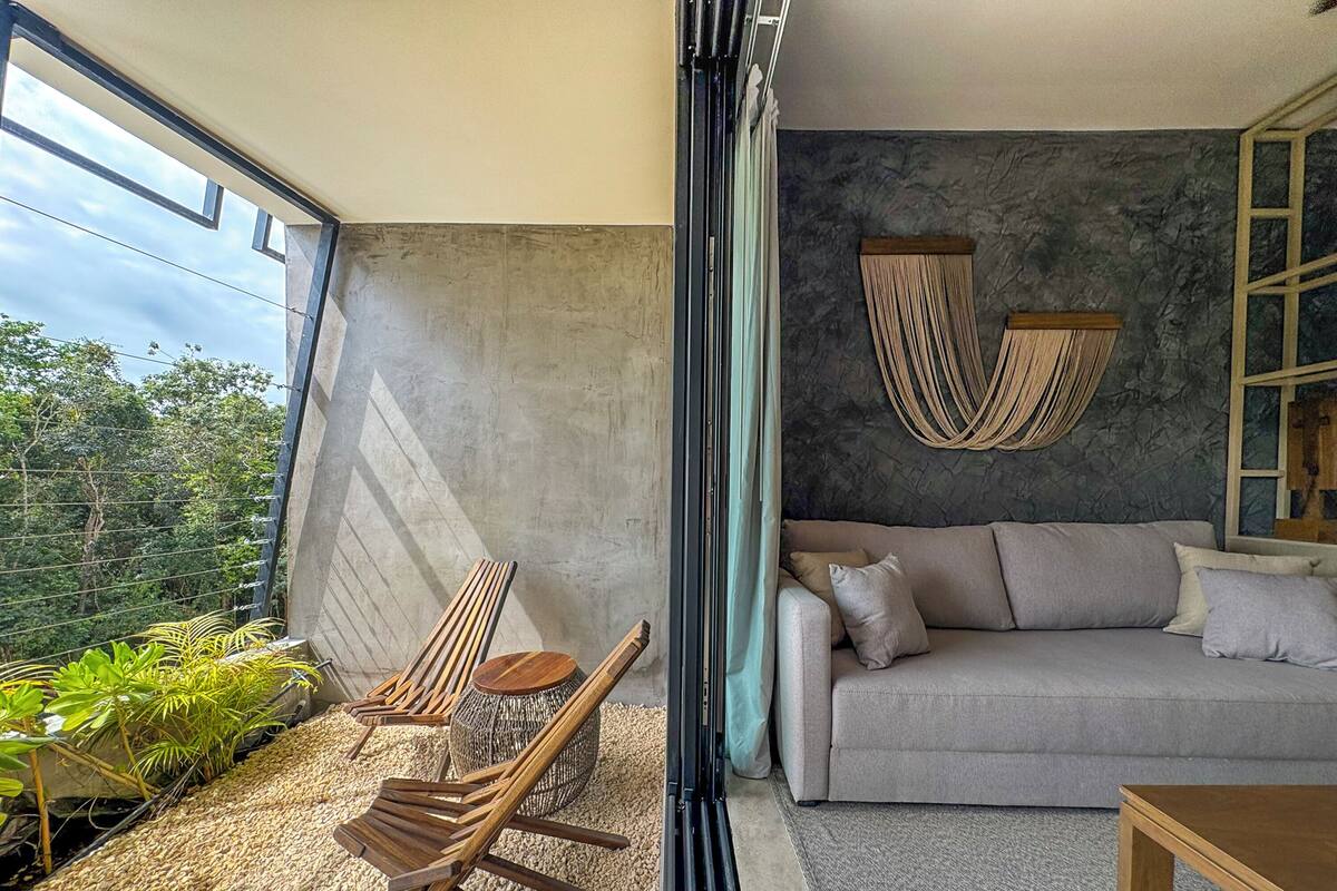 A modern living room with a grey sofa, wooden chairs, a coffee table, and a large window overlooking greenery.