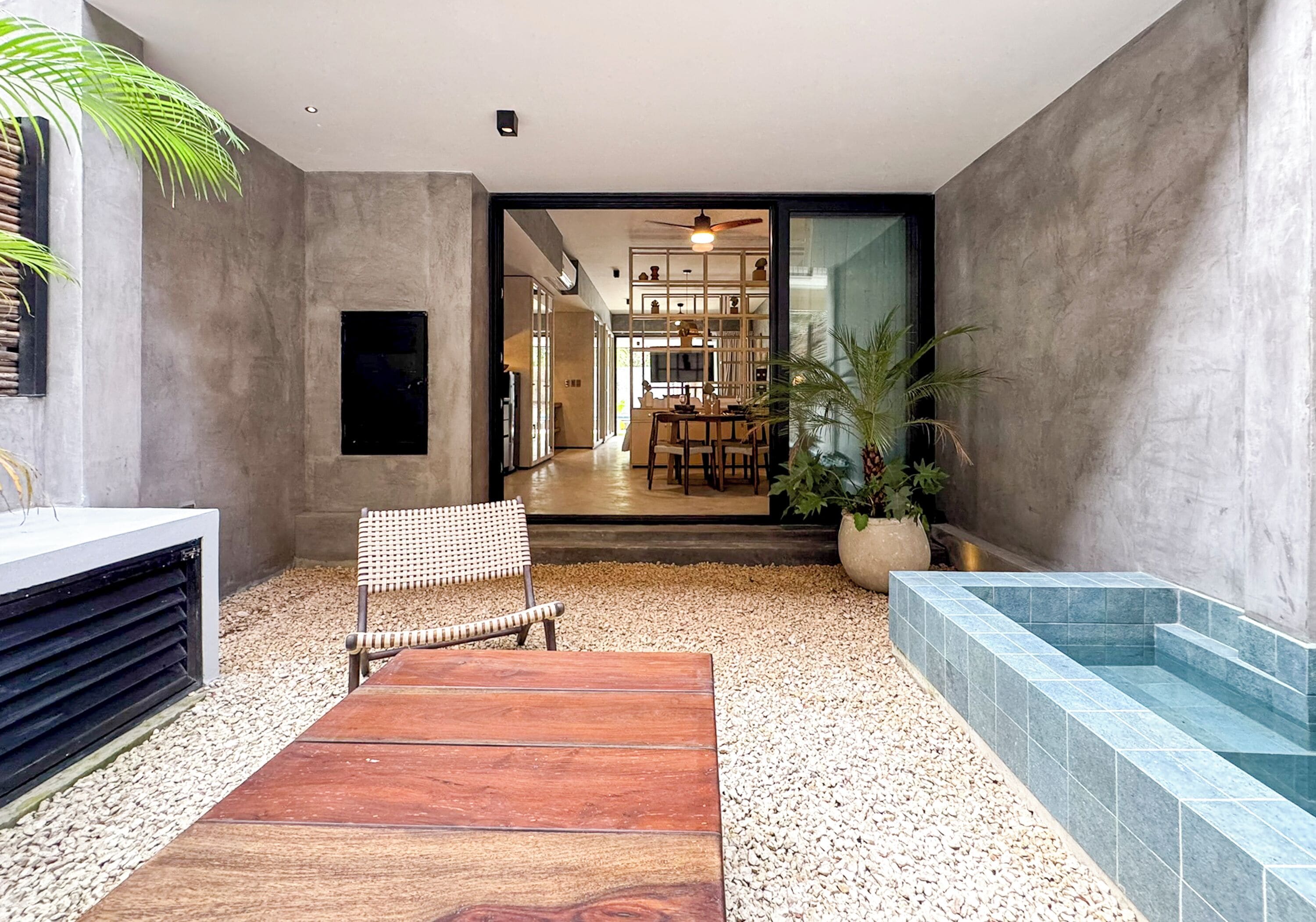 Un interior moderno con un banco de madera, una silla de mimbre, una planta en maceta y una zona de piscina con baldosas.
