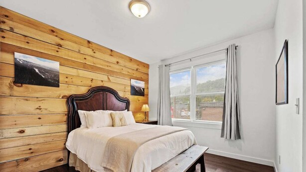 A bedroom with a wooden headboard, a bed with white bedding, a bench at the foot of the bed, a window with curtains, and two framed pictures on the wall.