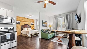 A compact hotel room with a kitchenette, a bed with a wooden headboard, a green sofa, and a dining table with chairs.