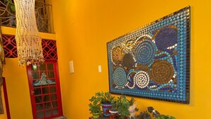 A hallway with a yellow wall, a mosaic artwork, a wooden table with potted plants, and a red door. - Casa Artística Pousada Leão (Maceio)