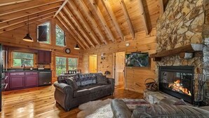 A log cabin interior with a stone fireplace, wooden ceiling beams, a kitchen area, and a cozy seating area. - Romantic Mountain view Cabin with Indoor/Outdoor Spa House + Sauna (Ellijay)