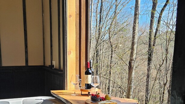 A hot tub with a wooden deck and a bottle of wine, surrounded by trees. - Romantic Mountain view Cabin with Indoor/Outdoor Spa House + Sauna (Ellijay)