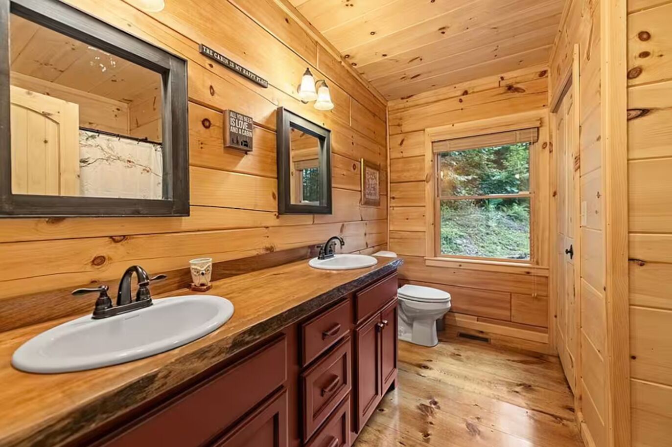 A wooden cabin bathroom with two sinks, a toilet, and a window providing a view of the outdoors.