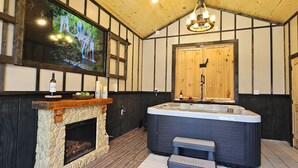 A room with a stone fireplace, a large hot tub, and a television mounted on the wall. - Romantic Mountain view Cabin with Indoor/Outdoor Spa House + Sauna (Ellijay)