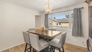 A dining room with a wooden table and four chairs, a chandelier, and a window with curtains.