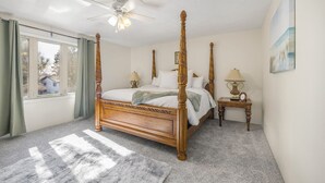 A four-poster bed with a wooden frame and a ceiling fan, a bedside table with a lamp, and a window with curtains.