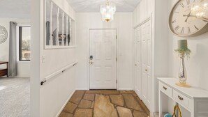 A hallway with a stone-patterned floor, white walls, a chandelier, a large wall clock, and a door.