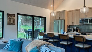 A modern kitchen with wooden cabinets, a stainless steel refrigerator, and a microwave. There is a dining area with a table and chairs, and a large window offering a view of the outdoors.