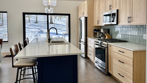 A modern kitchen with a large island, stainless steel appliances, wooden cabinets, and a view of a snowy landscape through the window.