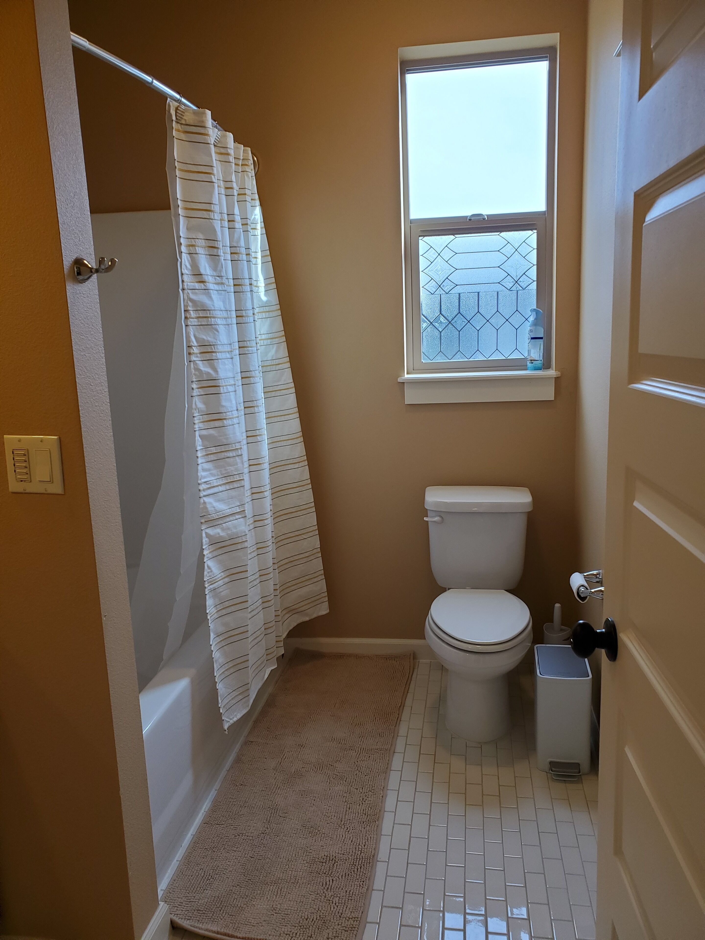 Une salle de bain avec des toilettes, une fenêtre ornée d’un motif géométrique, un rideau de douche et une petite poubelle.