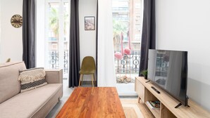Un salon comprenant un canapé, une table basse en bois, un téléviseur posé sur un meuble en bois, un fauteuil et un balcon doté d’une rambarde et de plantes.