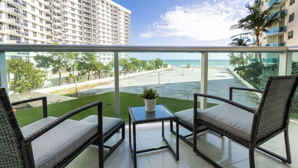 A balcony with wicker furniture, a small table with a plant, and a view of the ocean and buildings. - Ocean View Condo, Direct Beach Access, Beach Gear & Cleaning Included (Hollywood)