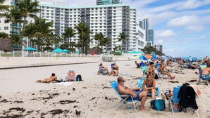 A beach with people sunbathing and sitting on beach chairs, palm trees, and a large hotel building in the background. - Ocean View Condo, Direct Beach Access, Beach Gear & Cleaning Included (Hollywood)