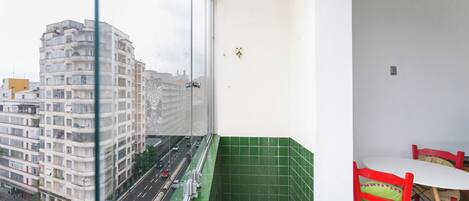 A balcony with a view of a city street, a green tiled wall, and a table with chairs.