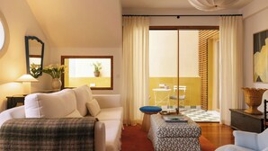 Hotellihuone, jossa on sänky, sohva, pieni pöytä lampun kanssa, tuoli, koristeellinen kori ja näkymä ulkopuolella olevalle parvekkeelle.