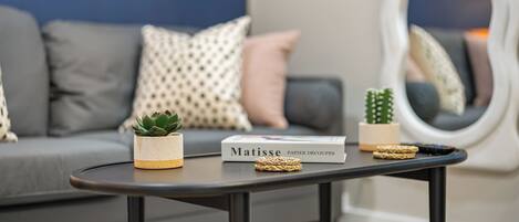 A modern living room with a grey sofa, patterned cushions, a small table with a book titled 'Matisse', a succulent plant, and a cactus.