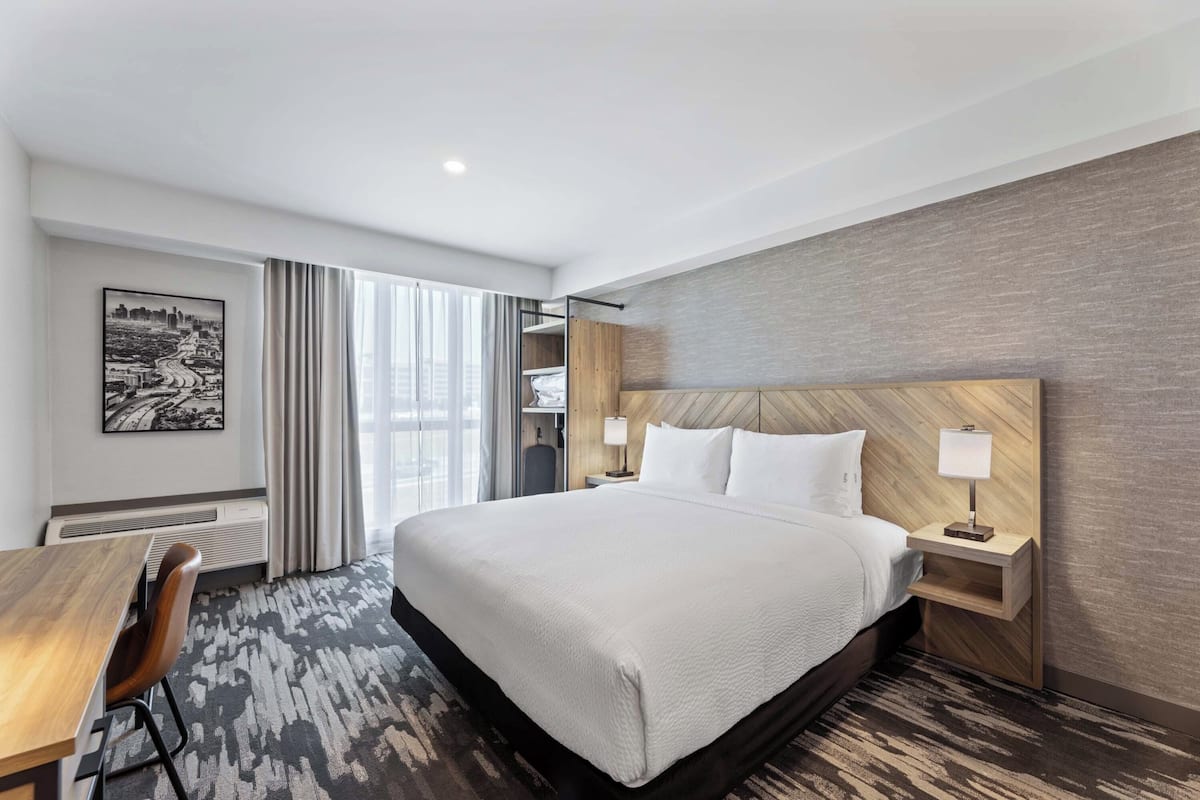A modern hotel room with a large bed, a desk, a chair, and a view of a cityscape through the window.
